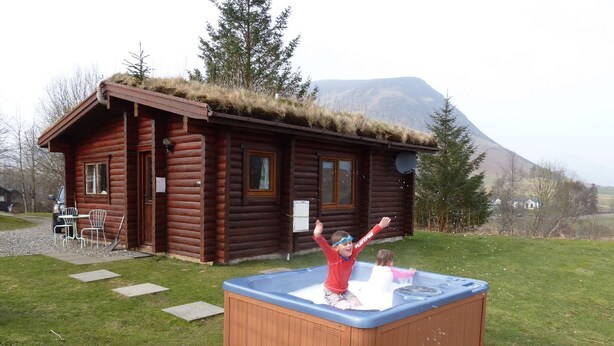 A log cabin with a hot tub in front, surrounded by a grassy area and a mountain in the background. - Log Cabin with Hot Tub for 2 | In the Cairngorm National Park | Accepts Dogs (Spittal of Glenshee)