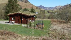 A rustic cabin with a thatched roof, a small porch, and a seating area with chairs. - Log Cabin with Hot Tub for 2 | In the Cairngorm National Park | Accepts Dogs (Spittal of Glenshee)