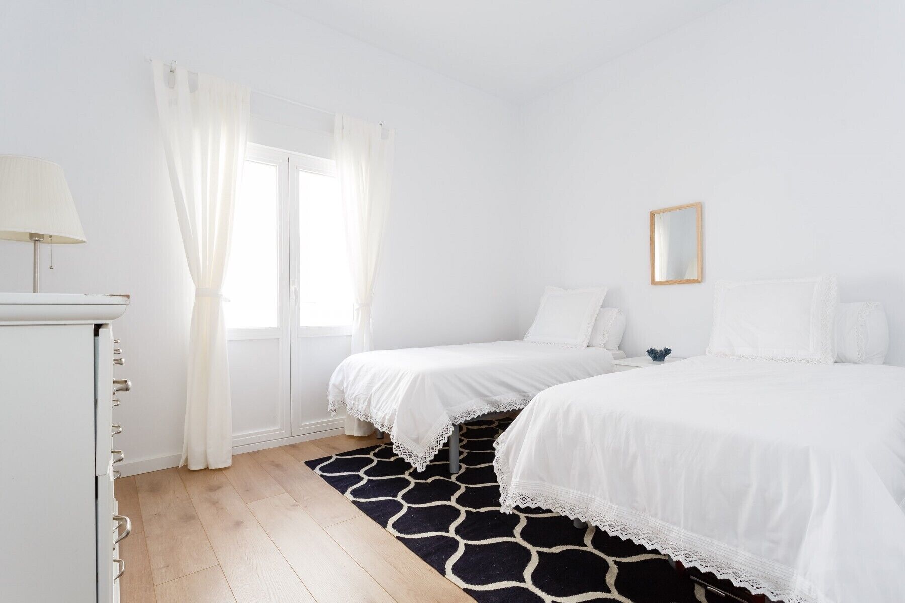 A bedroom with two beds, a window with white curtains, a bedside table with a lamp, and a black and white patterned rug.