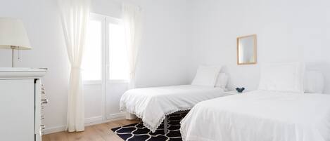 A bedroom with two beds, a window with white curtains, a bedside table with a lamp, and a black and white patterned rug.