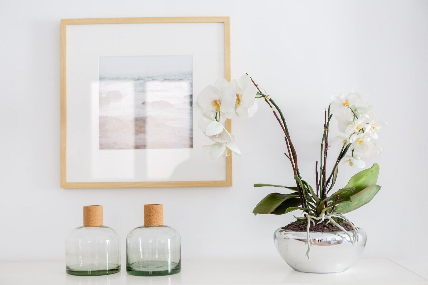 Un cuadro en la pared, una orquídea en maceta y dos botellas de vidrio con tapones de corcho.