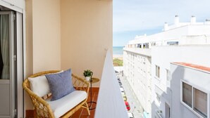 Een balkon met een rieten stoel, een kleine plant in pot en uitzicht op zee en omliggende gebouwen.