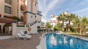 Ein Poolbereich mit Liegestühlen, einer Poolterrasse und einem Gebäude mit Balkonen und Palmen.