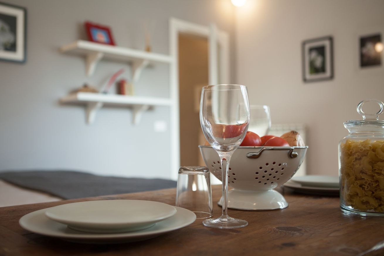A dining table with plates, a wine glass, a colander with tomatoes, and a jar of pasta.