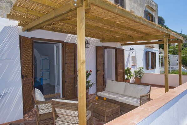 A covered outdoor seating area with wicker furniture and a sofa, a wooden pergola, and a view of the surrounding buildings. - Casa Sunset - Panoramic terrace overlooking Positano and Capri (Praiano)