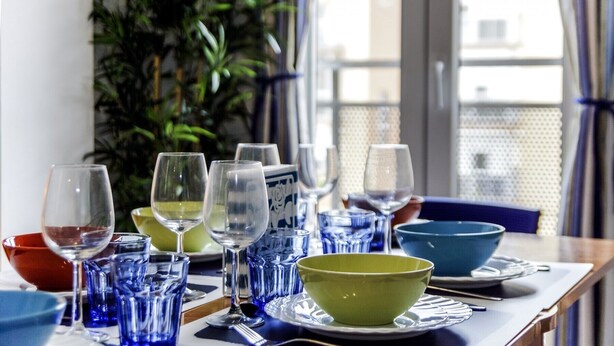 Mesa de comedor servida con diversos vasos y recipientes, una planta al fondo y vistas de edificios a través de una ventana.