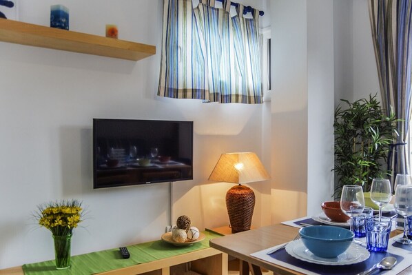 Un coin repas avec une table dressée pour deux personnes, un téléviseur fixé au mur, une lampe et une plante.
