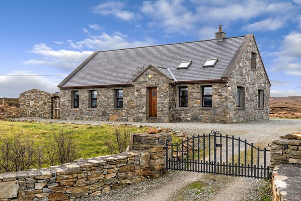 A stone house with a sloped roof, a wooden door, and a gravel driveway leading to a black metal gate. - Cottage 118 - Cleggan - sleeps 7 guests  in 3 bedrooms (Cleggan)