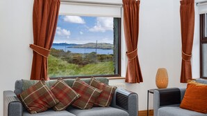 Una sala de estar con un sofá gris, cojines a cuadros, una mesa de centro de madera y vistas a un lago y colinas a través de una ventana con cortinas marrones.