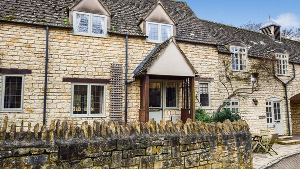 A stone building with a traditional design, featuring a central entrance with a sign, multiple windows, and a stone wall in front. - A sudeley castle cottage that sleeps 4 guests  in 2 bedrooms (Winchcombe)