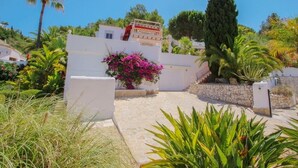 Un bâtiment blanc, avec un chemin de pierre menant à une entrée fermée par un portail, entouré d’une végétation luxuriante et de palmiers.