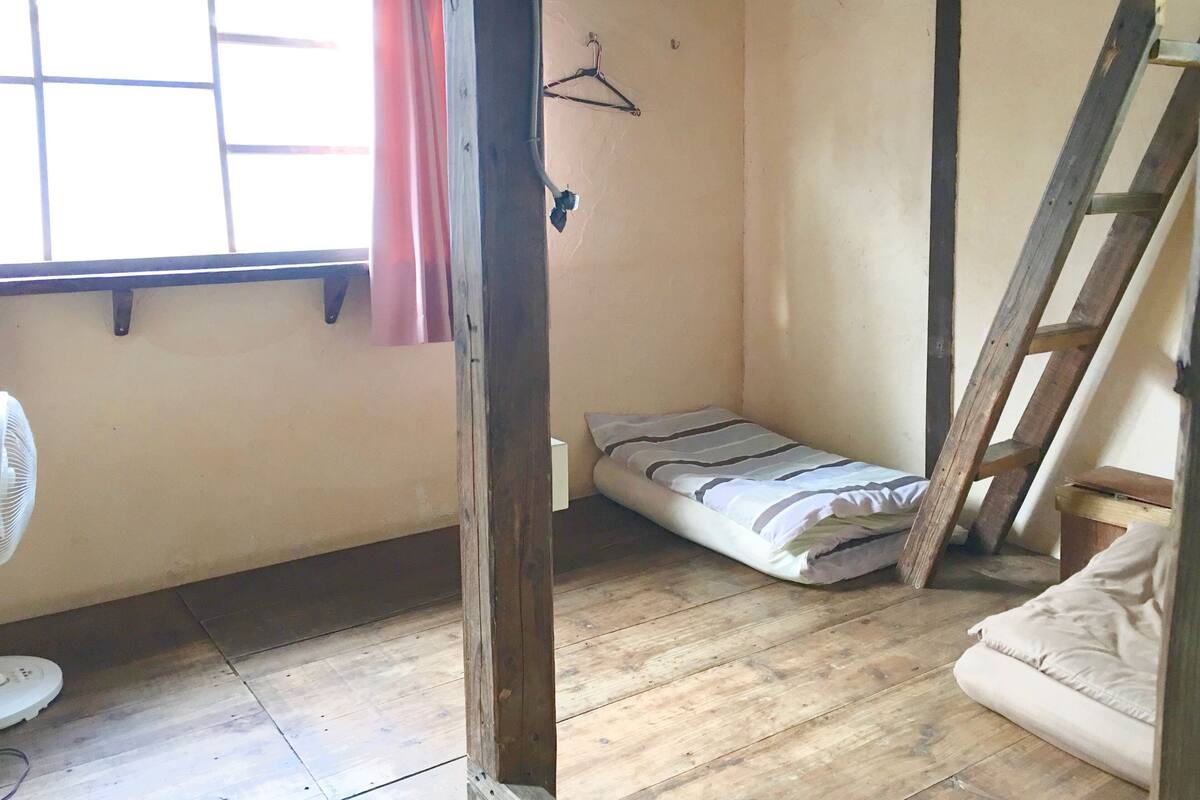 A small, single-room accommodation with a wooden floor, a bed, a fan, and a window with a curtain.