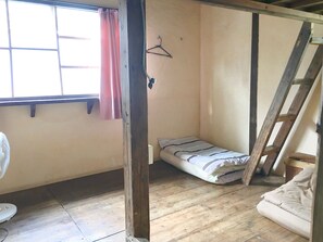 A small, single-room accommodation with a wooden floor, a bed, a fan, and a window with a curtain. - Gekkousou Okinawa - Hostel (Naha)