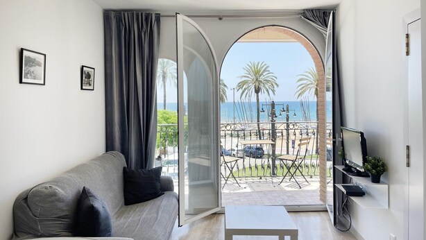 A modern living room with a grey sofa, a small white table, and a balcony with a view of the beach and palm trees. - RIBERA PETIT BY BLAUSITGES Small apartment with superb sea views in Sitges. (SITGES)