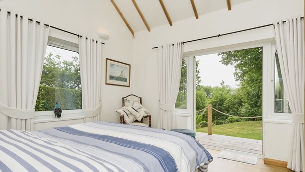 A bedroom with a large bed, two windows with curtains, a chair, and a view of greenery outside. - Kipper Cottage  -  a bungalow that sleeps 4 guests  in 2 bedrooms (Ryde)