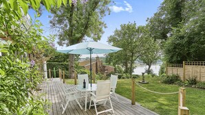 A wooden deck with a table and chairs under a large umbrella, surrounded by lush greenery and a view of a lake. - Kipper Cottage  -  a bungalow that sleeps 4 guests  in 2 bedrooms (Ryde)