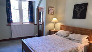 A bedroom with a wooden bed, floral bedding, a nightstand with a lamp, a window with curtains, a radiator, and a framed picture on the wall. - The Maid's House, Carmichael Country Cottages. 4 star comfortable cottage on rural Estate (Biggar)