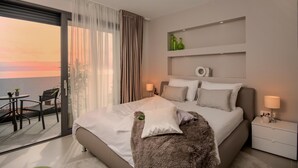 A modern hotel room with a large bed, a nightstand, a small table with chairs, and a balcony with a view of the sunset.