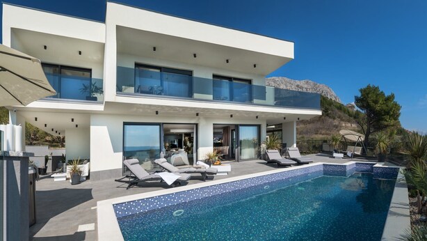 A modern two-story house with a swimming pool, outdoor seating, and a view of the mountains.