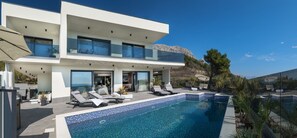 A modern two-story house with a swimming pool, outdoor seating, and a view of the mountains.