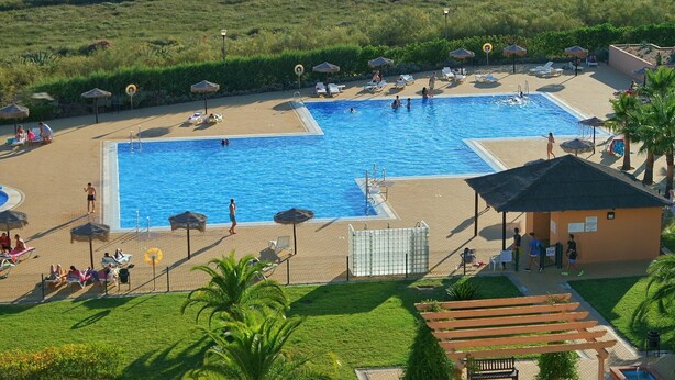 A large outdoor swimming pool with multiple lanes, surrounded by sun loungers and umbrellas, and a small building likely for pool maintenance or services.