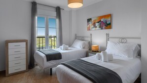 A hotel room with two beds, a wooden dresser, a window with curtains, and a painting on the wall.