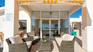 Un patio exterior con mobiliario de mimbre, una mesa de vidrio con una botella y un área de piscina con un cartel que enumera las normas de la piscina.