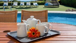 Una mesa de madera con una bandeja que contiene una tetera, tazas y una flor, con vista a una piscina con sillas de descanso y sombrillas.