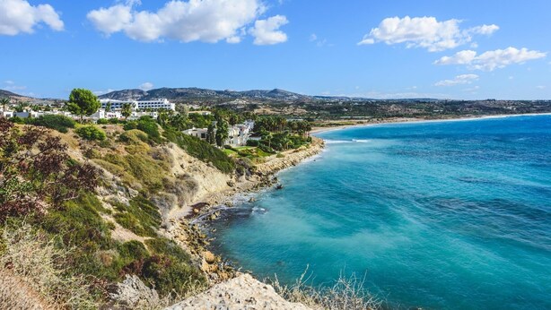 Un pueblo costero con edificios a lo largo de la orilla, un mar azul cristalino y un paisaje montañoso al fondo.