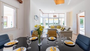 A modern dining area with a glass table, chairs, and a vase of flowers. There is a sofa and a dining table in the background with yellow cushions. - Villa Savoya Sunrise by Ezoria Villas (Coral Bay)