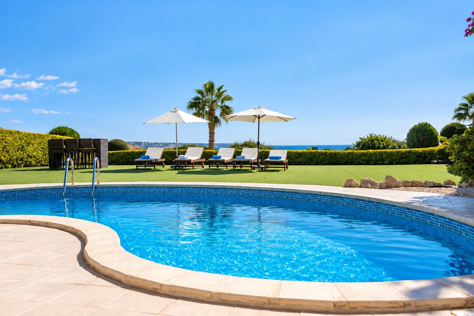 A swimming pool with lounge chairs and umbrellas, surrounded by a landscaped garden and a view of the sea.
