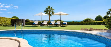 A swimming pool with lounge chairs and umbrellas, surrounded by a landscaped garden and a view of the sea.