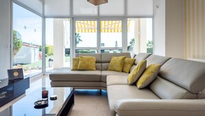 Una sala de estar moderna con un sofá seccional color beige, una mesa de centro de vidrio y una computadora portátil sobre un pedestal negro. Amplias ventanas ofrecen vistas a un jardín y zona de estar al aire libre.