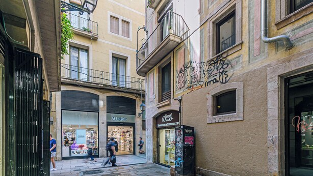 Una calle estrecha con edificios, una tienda cerrada llamada 'ALE-HOP' y una pequeña tienda con el letrero 'Vicenç 1775'.