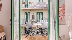 Un balcón con una mesa, sillas, una planta en maceta y vistas a un edificio con contraventanas verdes.