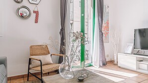 Una sala de estar con una silla de mimbre, un florero de vidrio con flores secas, una mesa de centro de madera y un televisor sobre un mueble.