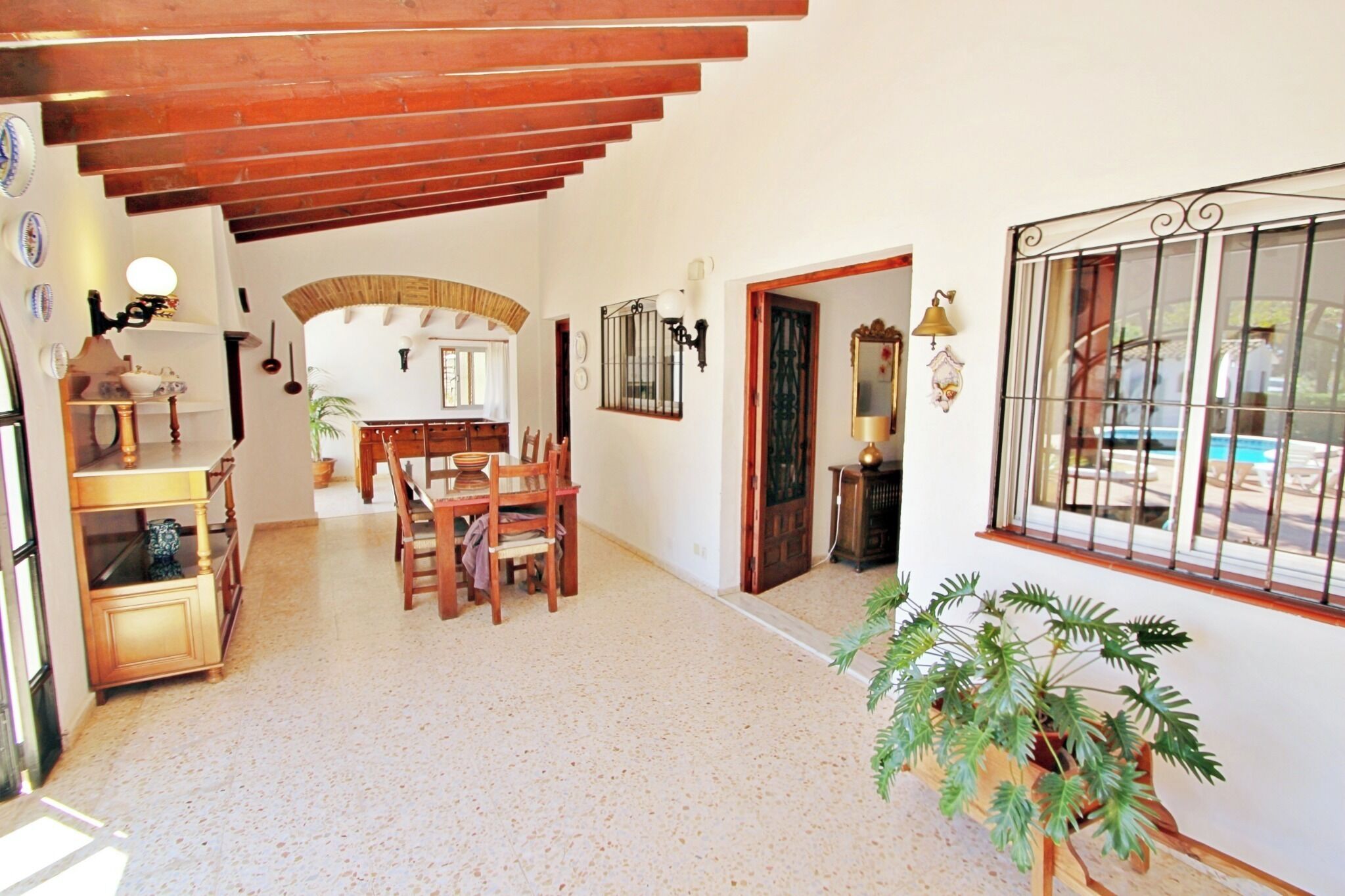 Amplia sala de estar con techo alto, vigas de madera, una mesa de comedor con sillas, una planta en maceta y vistas a través de una ventana con rejas.