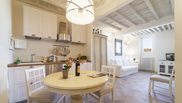 A kitchen with white cabinets, a dining table with chairs, a hanging light fixture, and a living area with a sofa and a television.