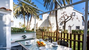 Une table à plateau en verre, sur laquelle sont disposés des bols blancs, un panier de fruits et des couverts, le tout placé devant un mur blanc, avec une clôture en bois et des palmiers en arrière-plan.