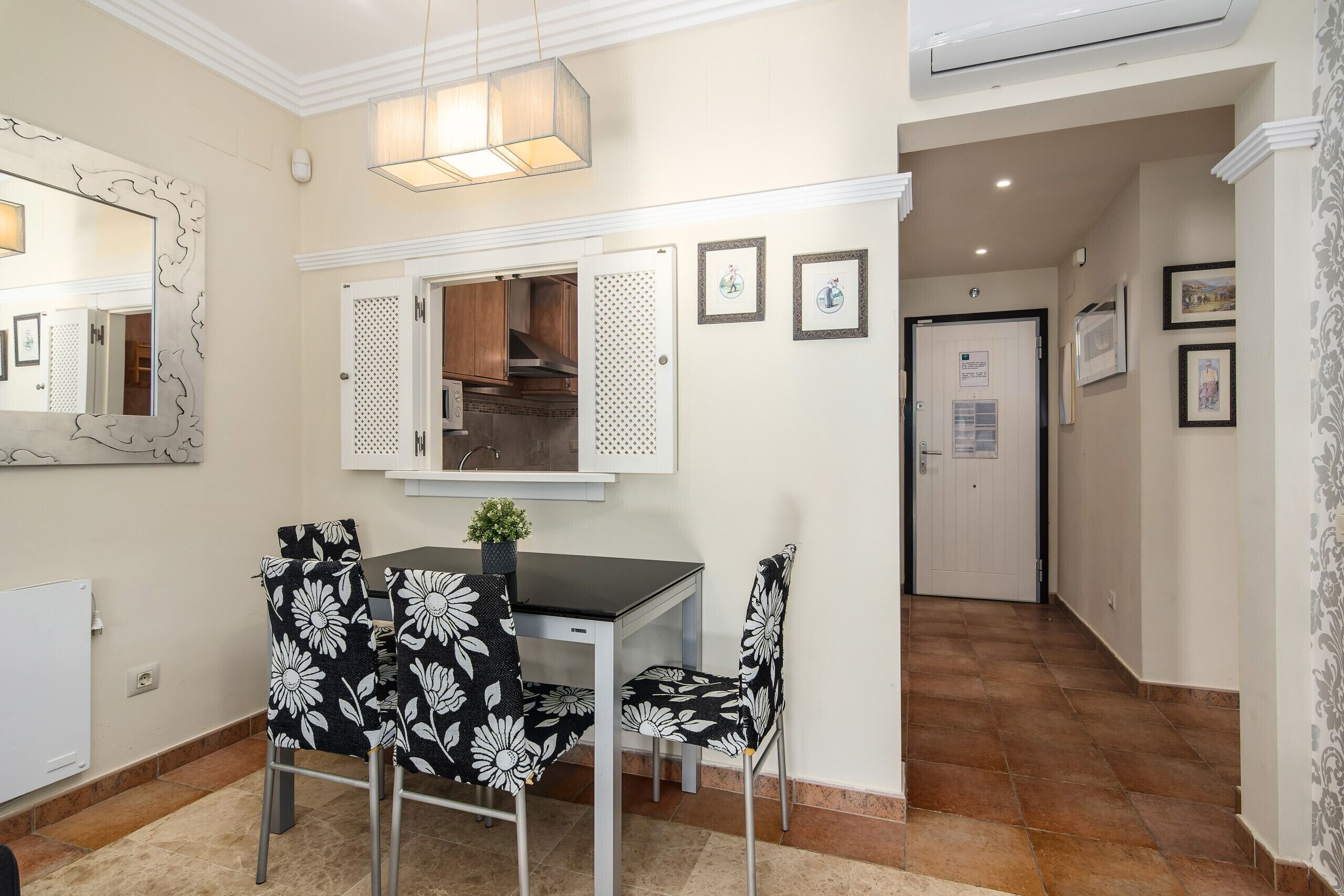 A dining area with a table and chairs, a mirror, and a kitchen visible through a window.