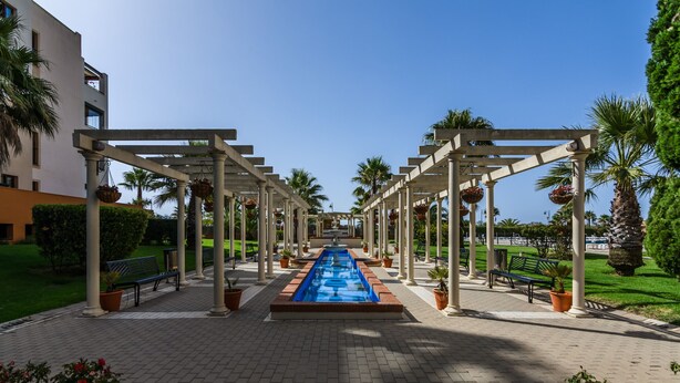 Zona de piscina con una fuente central, rodeada de pérgolas blancas y plantas en macetas.