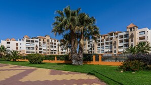 Un edificio residencial de varios pisos con balcones, una palmera en el centro y un camino pavimentado.