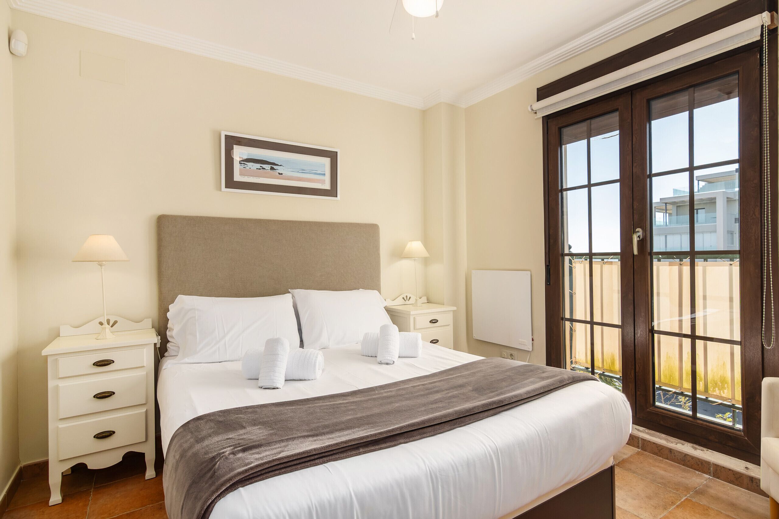 A hotel room with a bed, two bedside tables, a nightstand, a window with a view, and a framed picture on the wall.