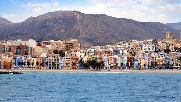A coastal cityscape with colorful buildings, a beach, and mountains in the background. - HAPPY  - ESTUDIO VISTAS A CALLE (Villajoyosa)