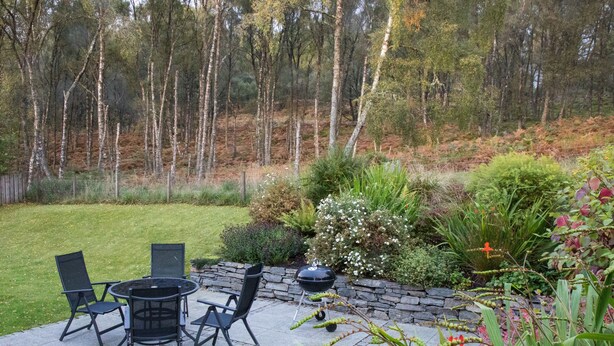 A patio with a round table and four chairs, surrounded by a stone wall and various plants. - Highland Holiday Homes - Tulloch Ard (Aviemore)