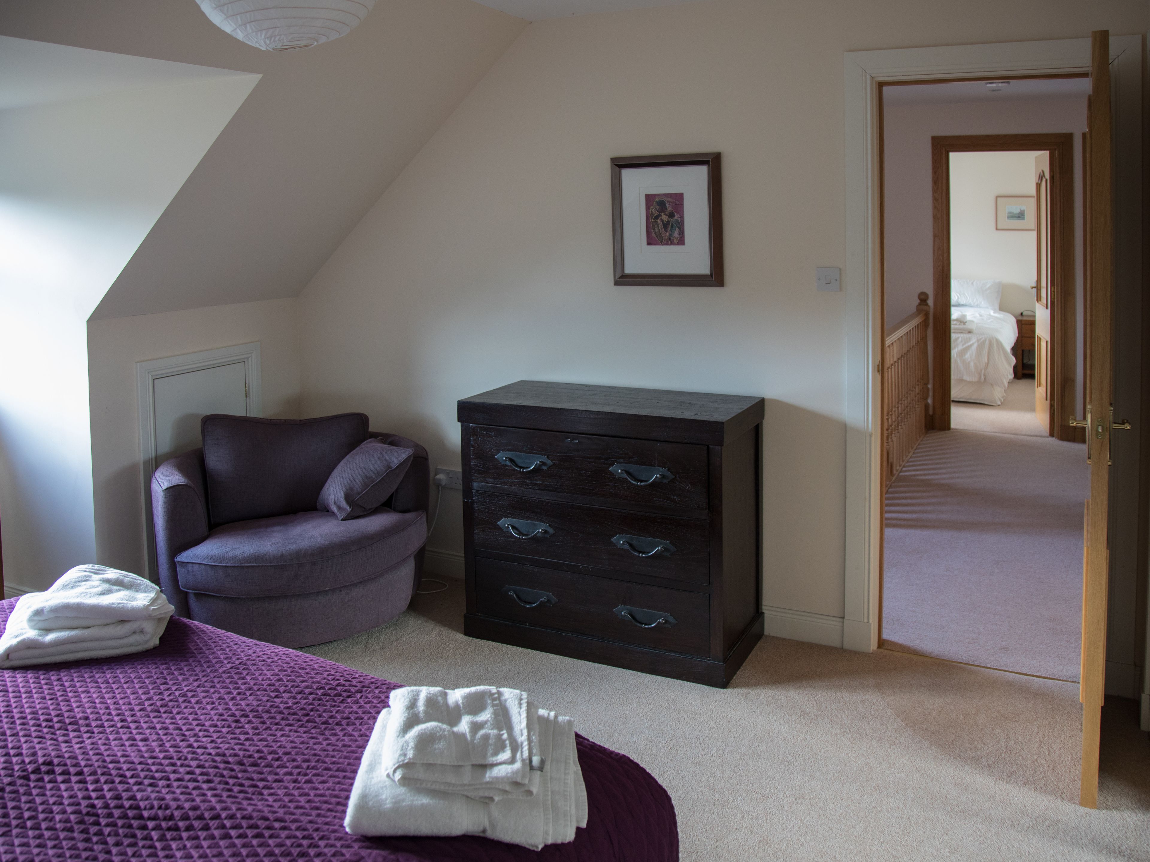 A bedroom with a purple bed, a dark dresser with drawers, a chair, and a framed picture on the wall.