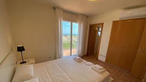 Ein Schlafzimmer mit Bett, Nachttisch, Lampe, Kleiderschrank und Blick ins Freie.