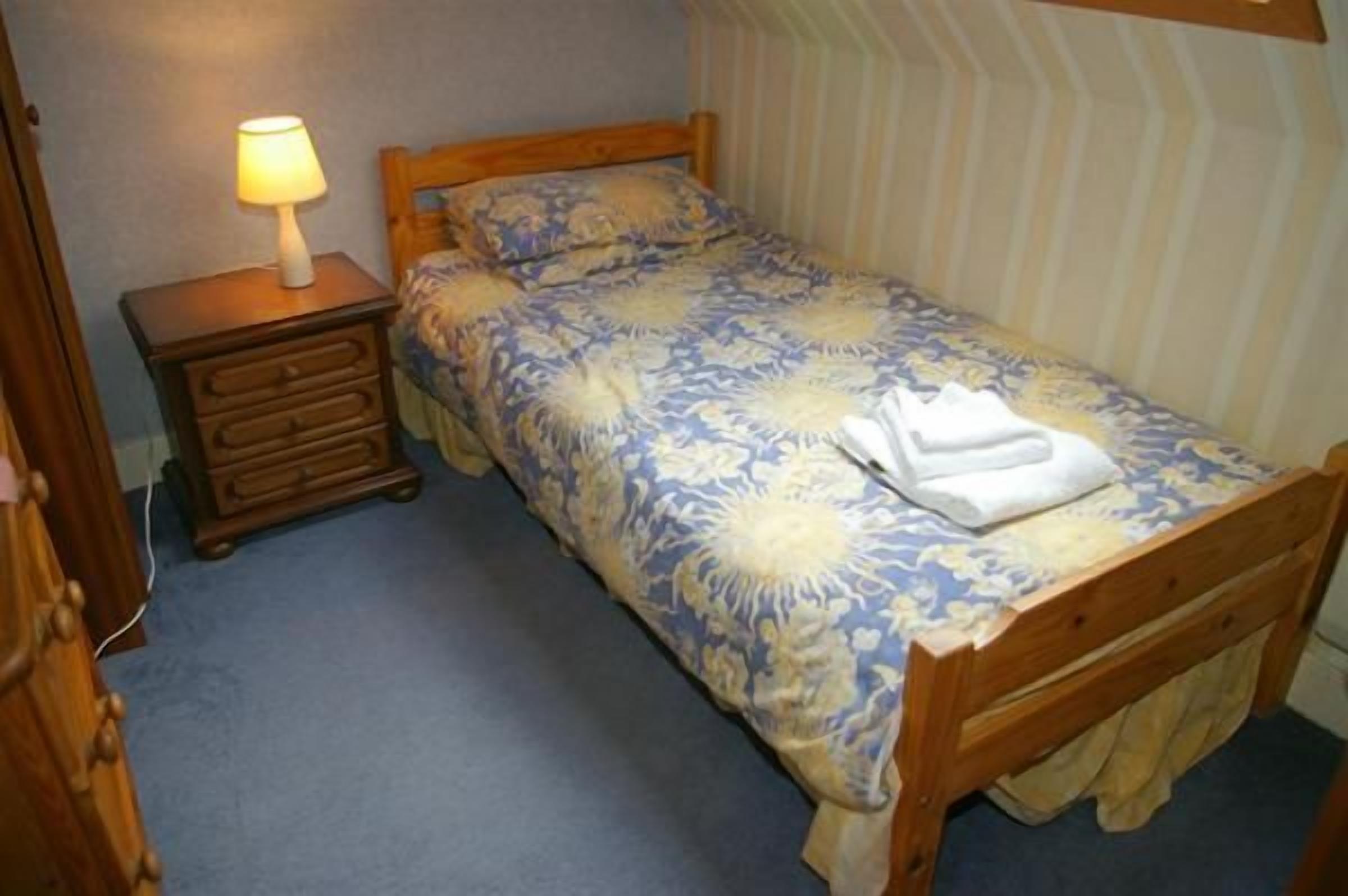 Een enkel bed met een bloemenprint deken, een houten nachtkastje met een lamp, en een gestreepte wand.