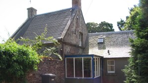 Un edificio de piedra con techo a dos aguas, una pequeña estructura adosada con detalles azules y un contenedor de basura negro.