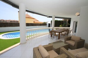 Eine überdachte Terrasse mit Pool, Gartenmöbeln und Blick auf ein Gebäude mit Ziegeldach.